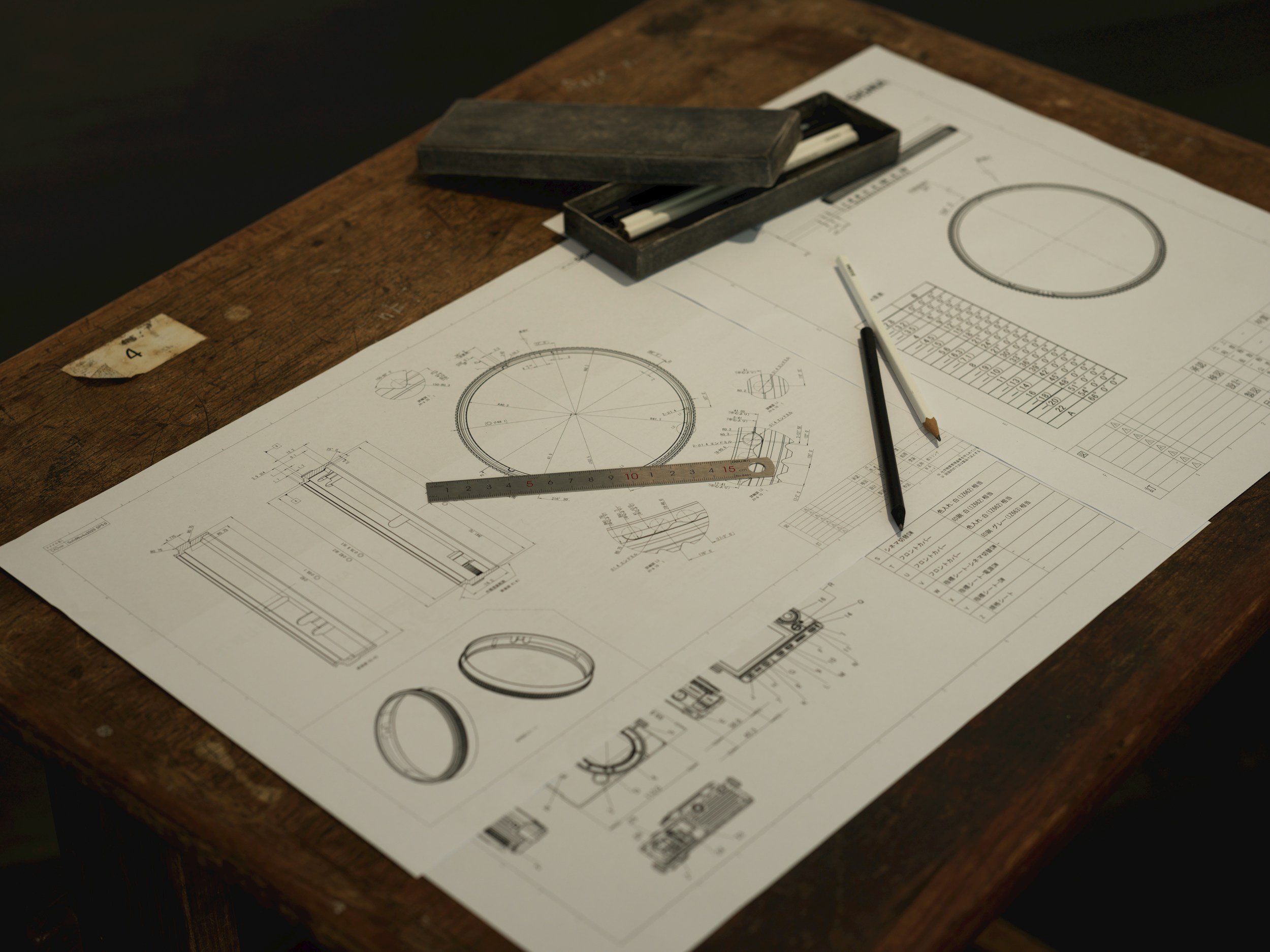 Detailed engineering drawings laid out on a wooden workbench with a steel ruler, a mechanical pencil and a drafting instrument case.