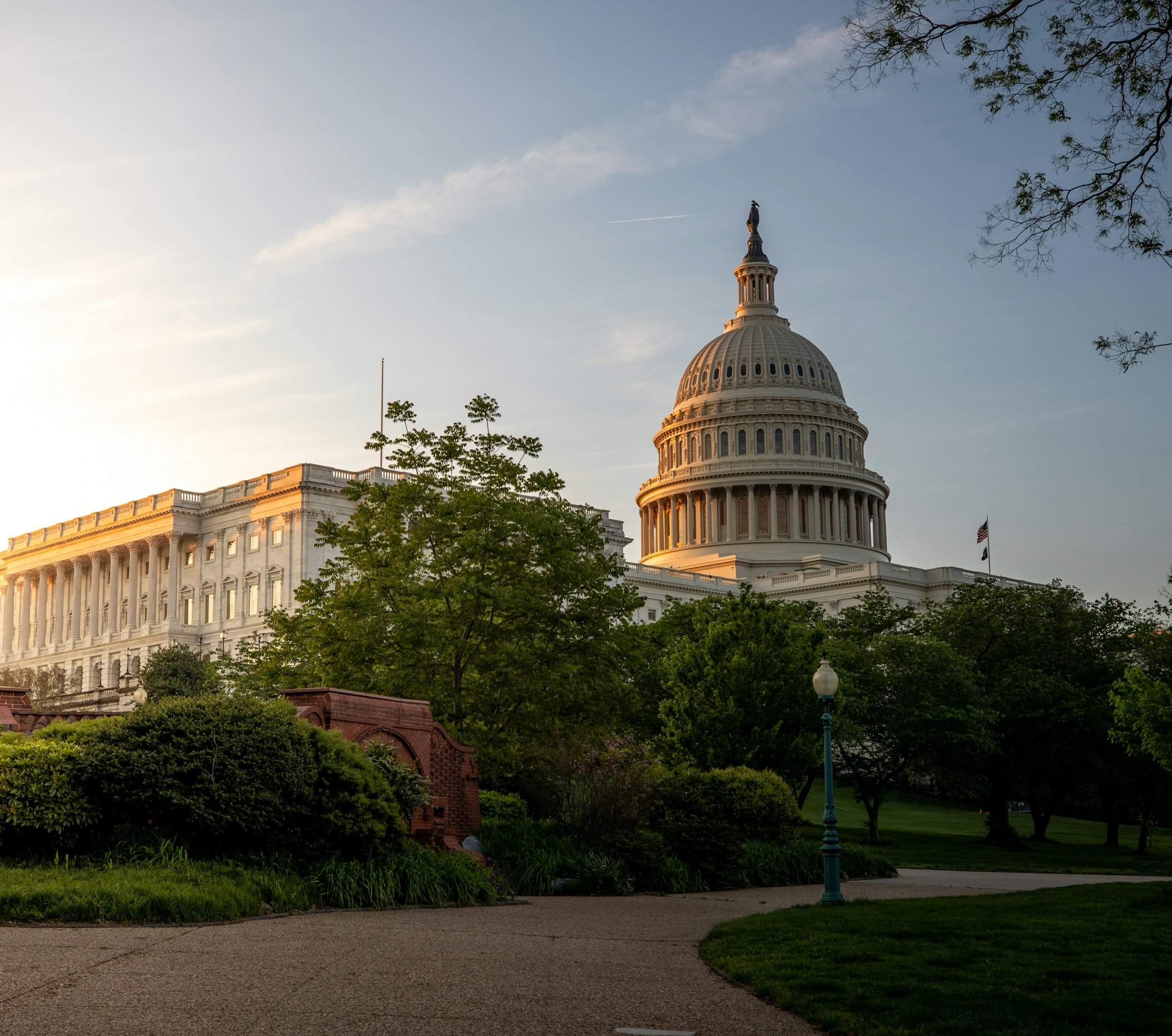 The United States Capitol building at sunrise, its dome lit by low sun and framed by trees and lawn.